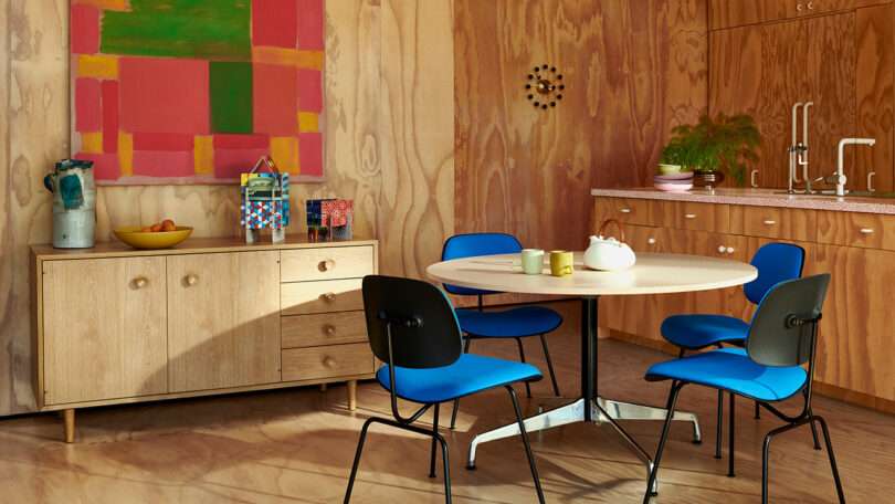 A kitchen with wood-paneled walls, blue chairs around a round table with a teapot and mugs, a sideboard, and a colorful abstract painting.