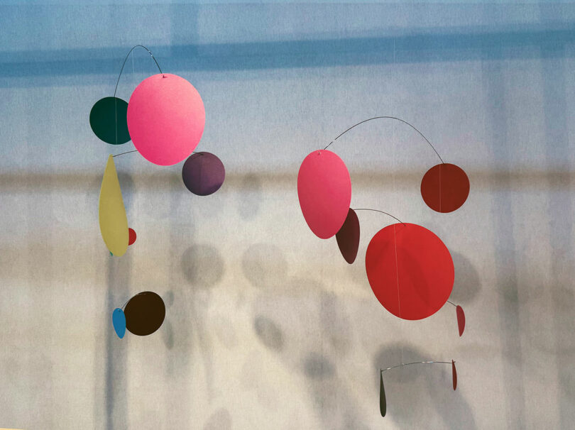 Two abstract mobiles made of colorful, geometric shapes hang against a neutral wall, casting soft shadows—evoking the playful elegance often seen at Maison & Objet.