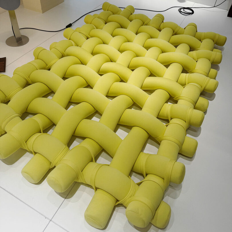 A woven, padded, bright yellow mat with thick, interlacing cylindrical cushions on a white floor; a power cord and part of a lamp are visible in the background, evoking the vibrant design spirit of Maison & Objet.