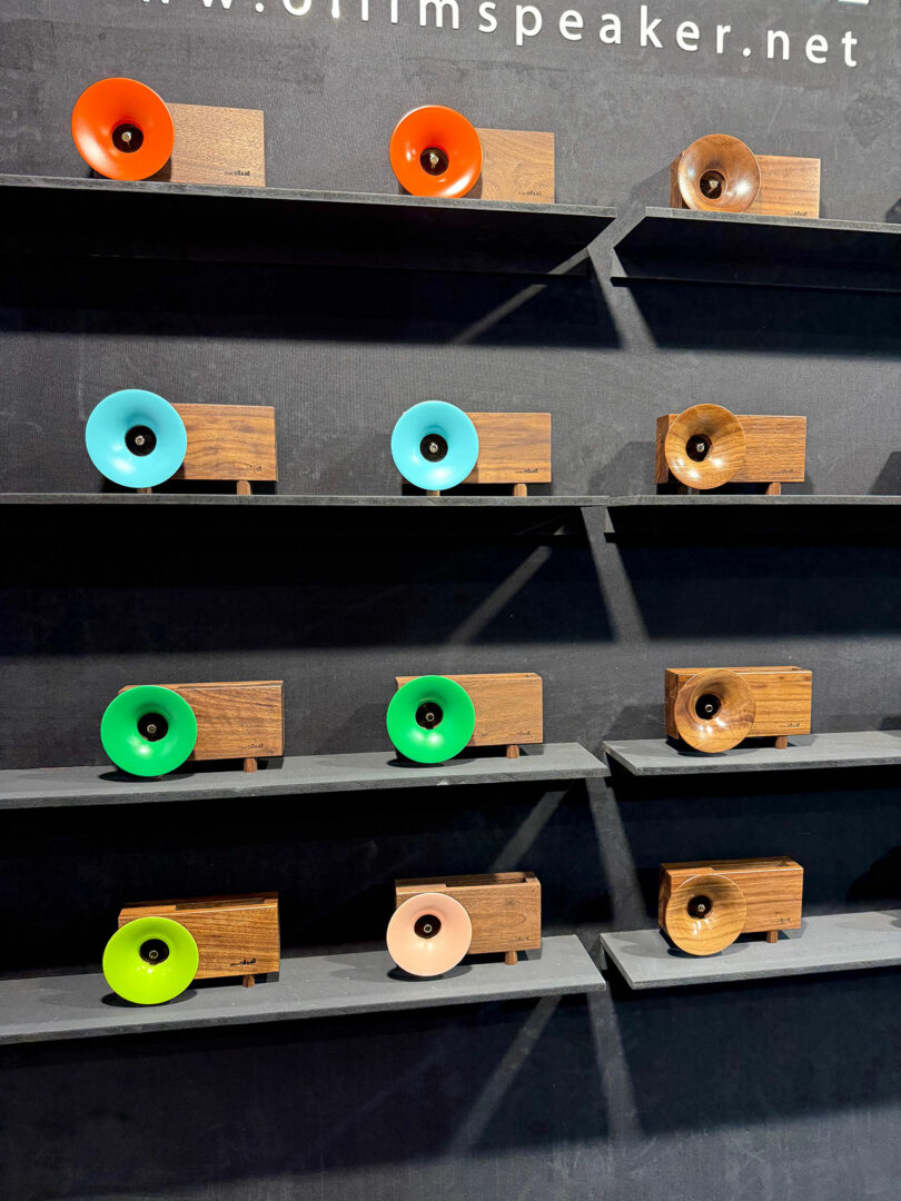 Wooden speakers with colorful cone fronts in blue, orange, green, brown, and yellow are displayed on three black shelves against a dark background at Maison & Objet.