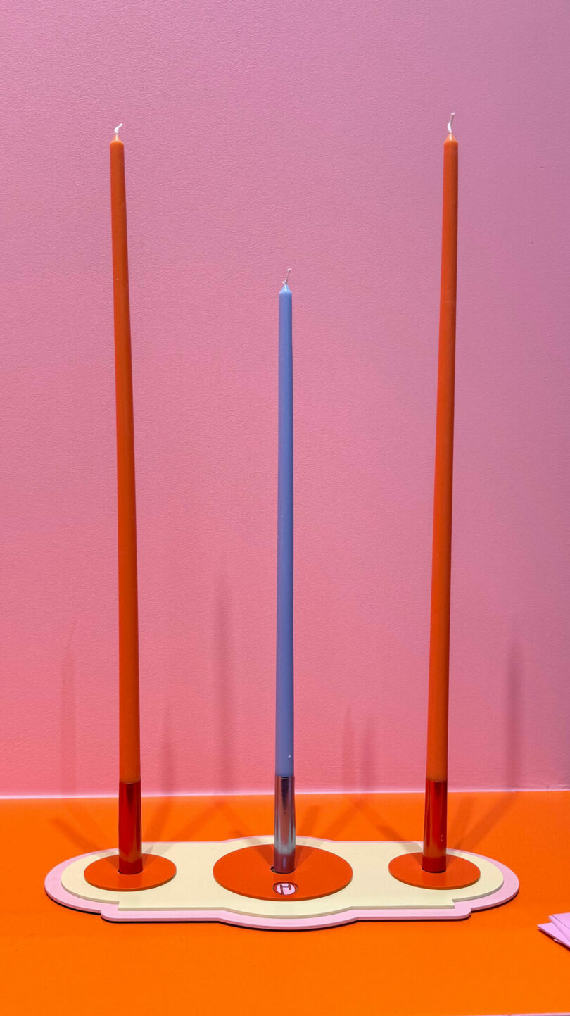 Three tall candles—two orange and one blue—stand upright in a modern, flat candle holder against a pink and orange background, evoking the chic style often seen at Maison & Objet.