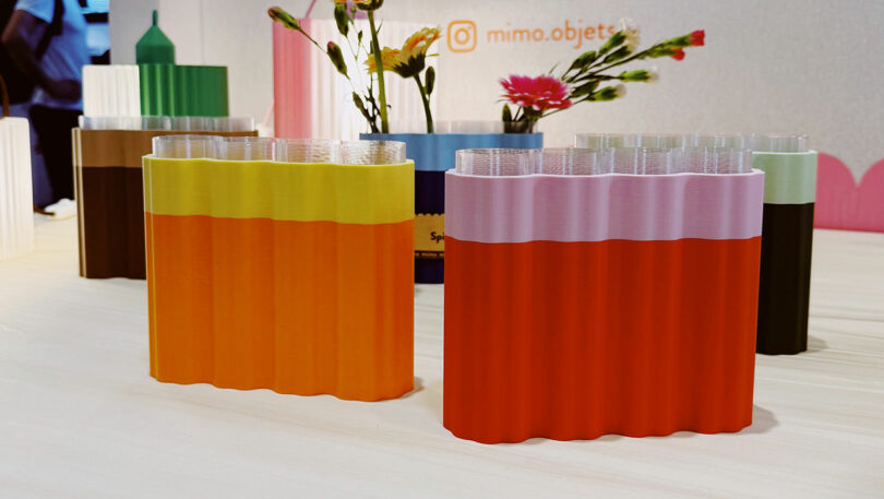 Colorful, wavy-edged vases with glass inserts hold flowers on a light table; more vases and an Instagram handle, 