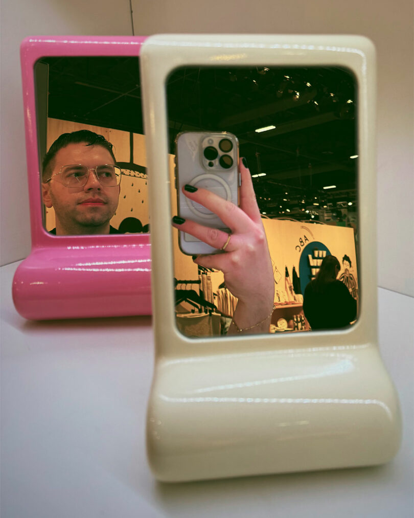 A person snaps a photo with a phone in front of a cream-colored mirror, while a man’s reflection appears in a separate pink Maison & Objet mirror behind it.