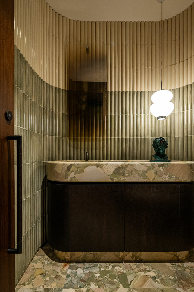 A bathroom features vertically ribbed beige and green tiles, a dark wood vanity with a stone countertop, a round white pendant light, and a small decorative object.