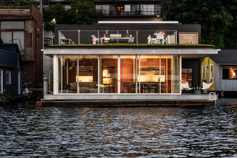 Modern floating house with large glass windows, rooftop seating area, and interior lights on, situated on a body of water with trees and buildings in the background.