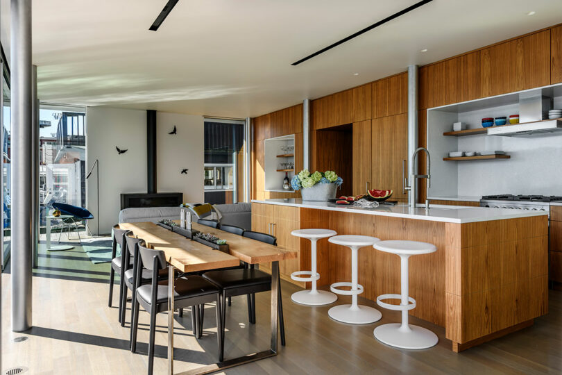 Modern open-plan kitchen and dining area with wooden cabinets, a large island with white bar stools, a wooden dining table, and lounge area in the background.