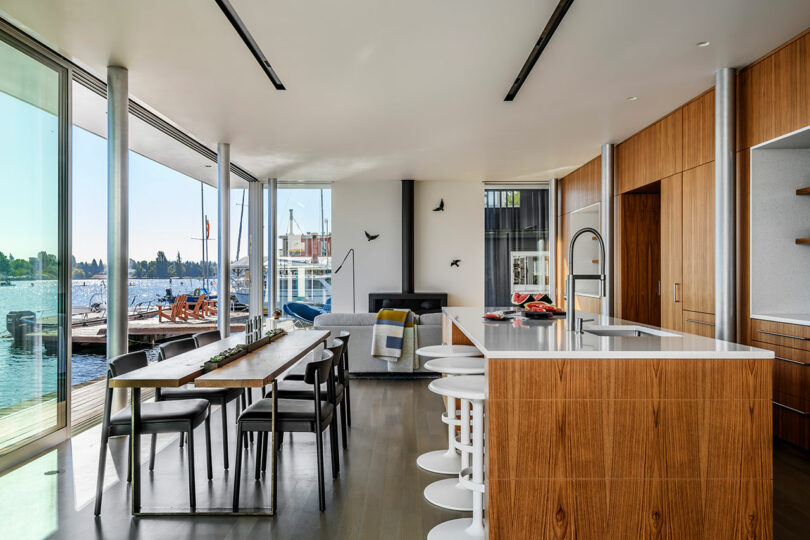 Modern open-concept kitchen and dining area with wood finishes, large windows overlooking a waterfront, and minimalist furnishings.
