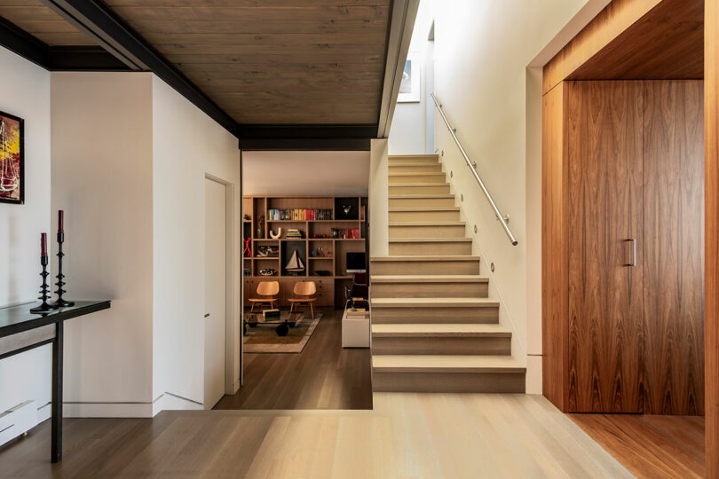Modern interior with a wooden staircase, white walls, built-in wooden storage, and an adjacent study room with bookshelves and mid-century style furniture.