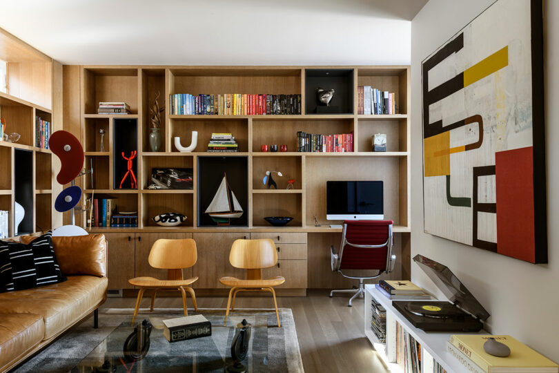 Modern home office with wooden bookshelves, colorful books, an iMac on a desk, two wooden chairs, abstract art, a record player, and a brown leather sofa.
