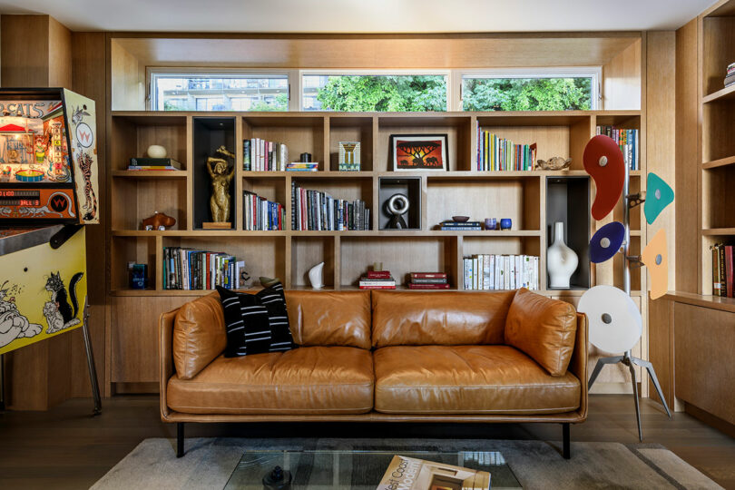 A tan leather sofa sits in front of built-in wooden bookshelves filled with books, art objects, and decor, with a pinball machine on the left and modern sculptures on the right.