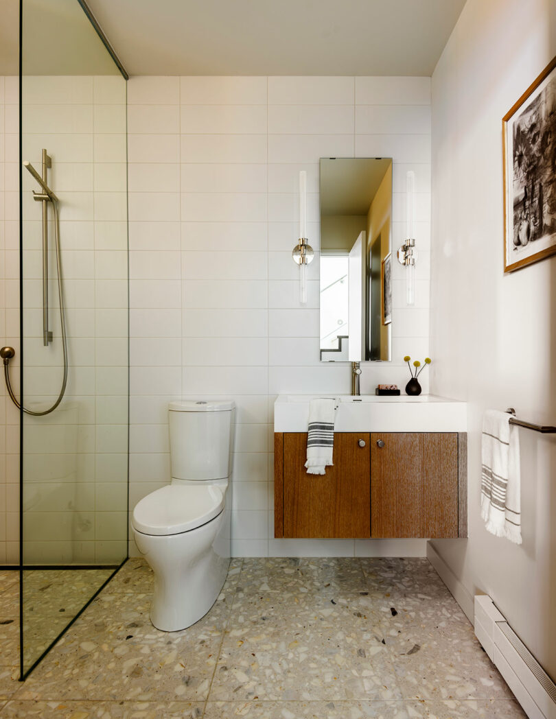 Modern bathroom with a white toilet, wood vanity, rectangular mirror, glass shower enclosure, and white tiled walls. A towel hangs on the sink and art is mounted on the wall.