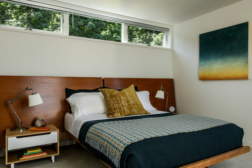 A modern bedroom with a wooden bed, blue bedding, a yellow accent pillow, two bedside tables with lamps, and a large abstract painting on the wall.