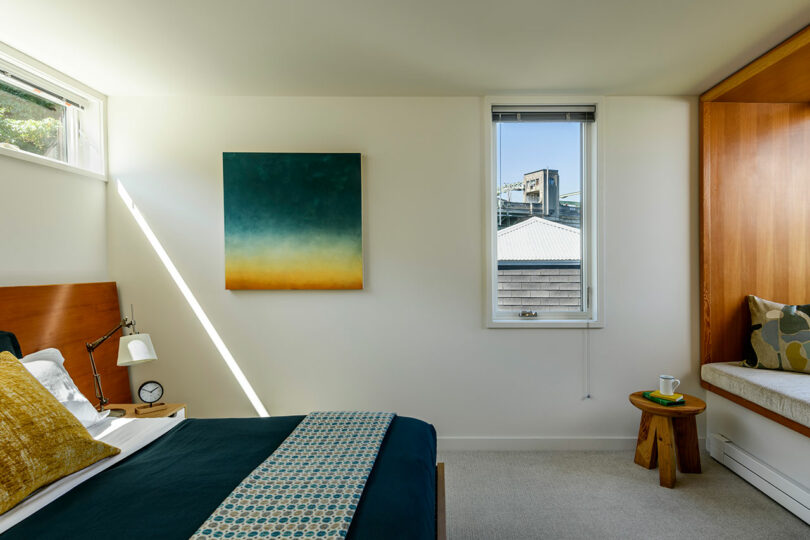 A modern bedroom with a wooden bed, blue and yellow bedding, a large abstract painting, a window seat with cushions, and sunlight streaming through a window.