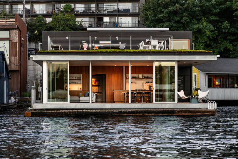 A modern houseboat with large glass windows and a rooftop deck is docked on a body of water, with neighboring buildings and greenery in the background.