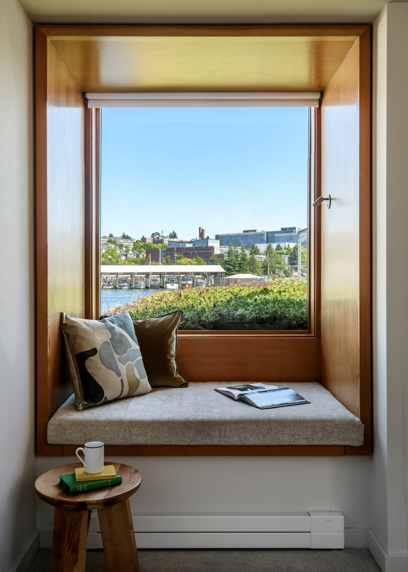 A built-in window seat with two pillows and an open book overlooks a waterfront view; a small side table holds a cup and two books.