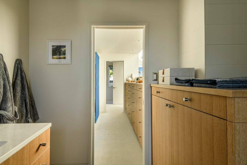 View of a hallway with built-in wooden cabinets and drawers; two towels hang on the left wall, with framed art above them, and natural light fills the space from windows ahead.