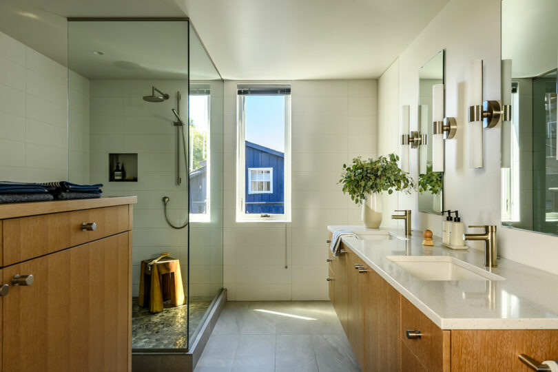 Modern bathroom with glass shower, wood cabinets, dual sink vanity, large mirror, potted plant, and a window letting in natural light.