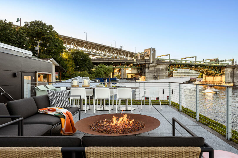 Modern outdoor patio with a fire pit, black sofa, bar seating, and a dining area overlooking a river and a nearby bridge under clear skies.