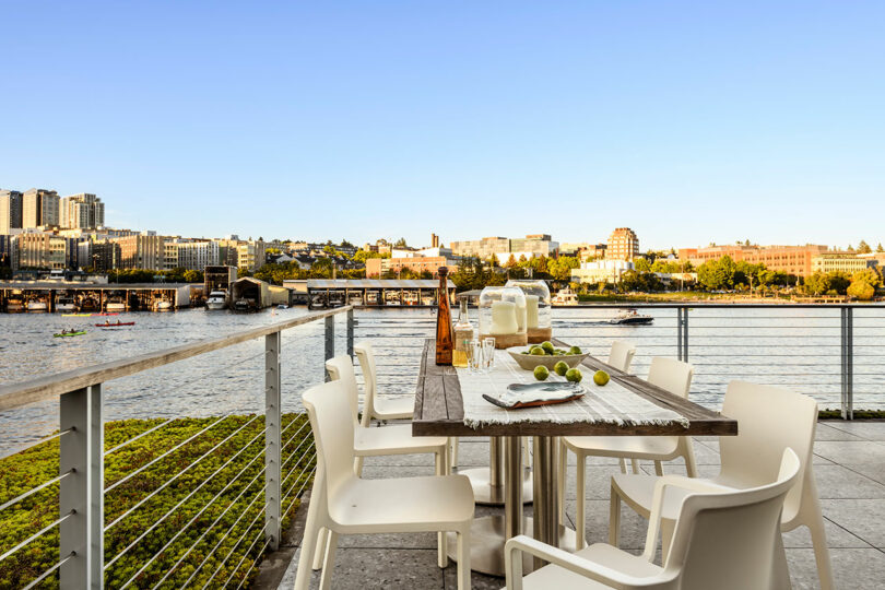 Outdoor dining table set with white chairs, glassware, and limes on a deck overlooking a calm river and cityscape under a clear blue sky.