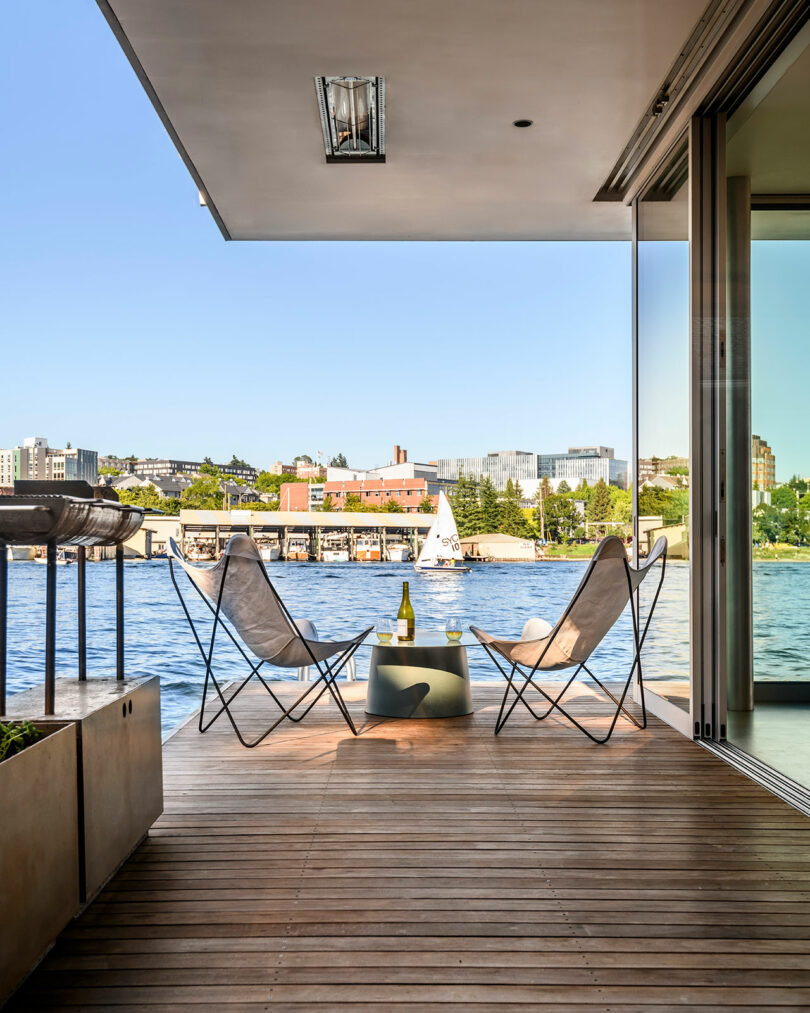 A waterfront deck with two chairs and a small table holding a bottle and glass, overlooking a calm body of water and city buildings under a clear sky.