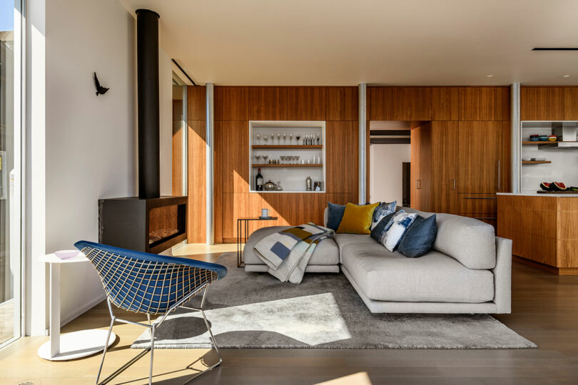 Modern living room with wood-paneled walls, a gray sectional sofa with colorful pillows, a gray rug, a black wood stove, and a blue accent chair near large windows.