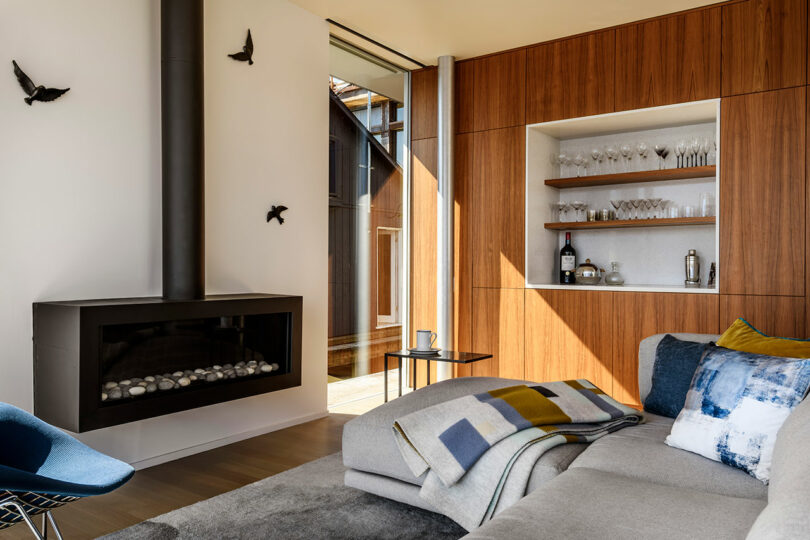 Modern living room with a black fireplace, bird wall decor, built-in wooden bar area with glassware, and a gray sectional sofa with a blanket and pillows.