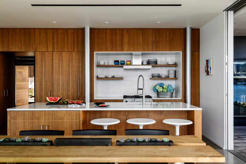 Modern kitchen with wood cabinets, white countertops, open shelving, and barstools; a dining table with succulents is in the foreground.