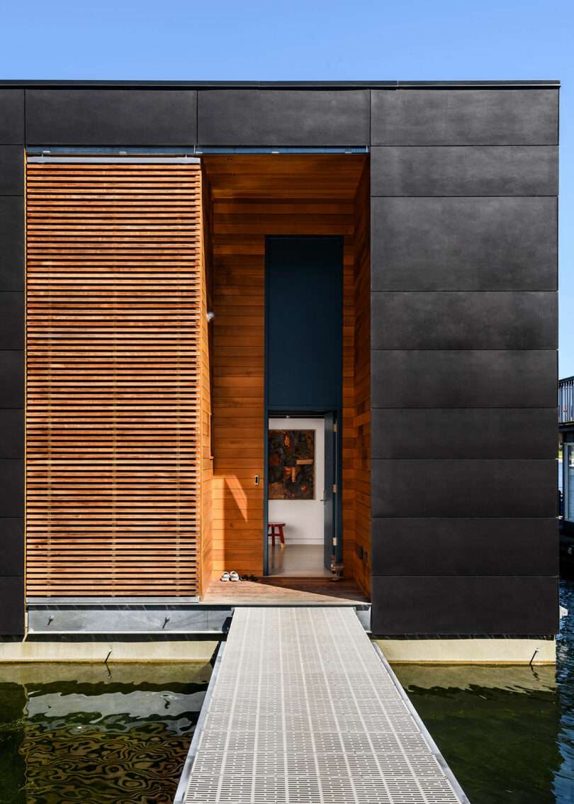 Modern floating house with dark exterior panels and wooden slats, viewed from a walkway leading to an open entryway over the water.