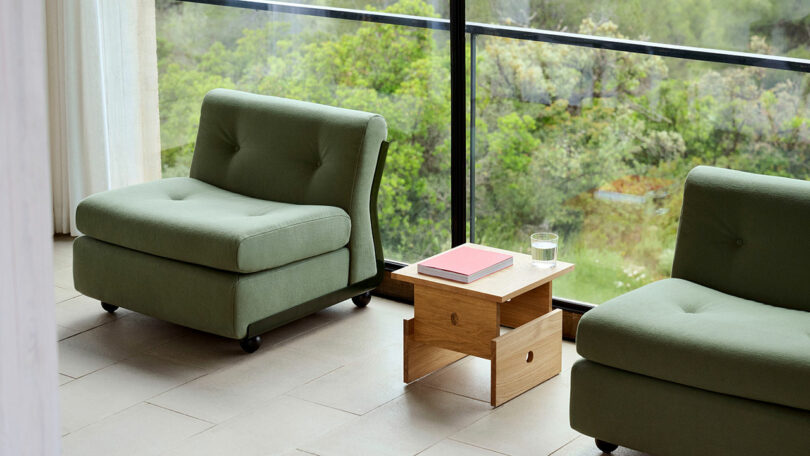 Two green armchairs are positioned by a large window with a view of greenery; a small wooden table with a pink book and a glass of water sits between them.