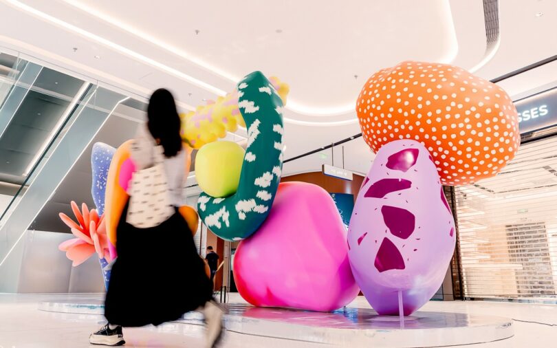 A person walks past large, colorful Studio Proba abstract sculptures resembling oversized mushrooms and plants in a brightly lit indoor space