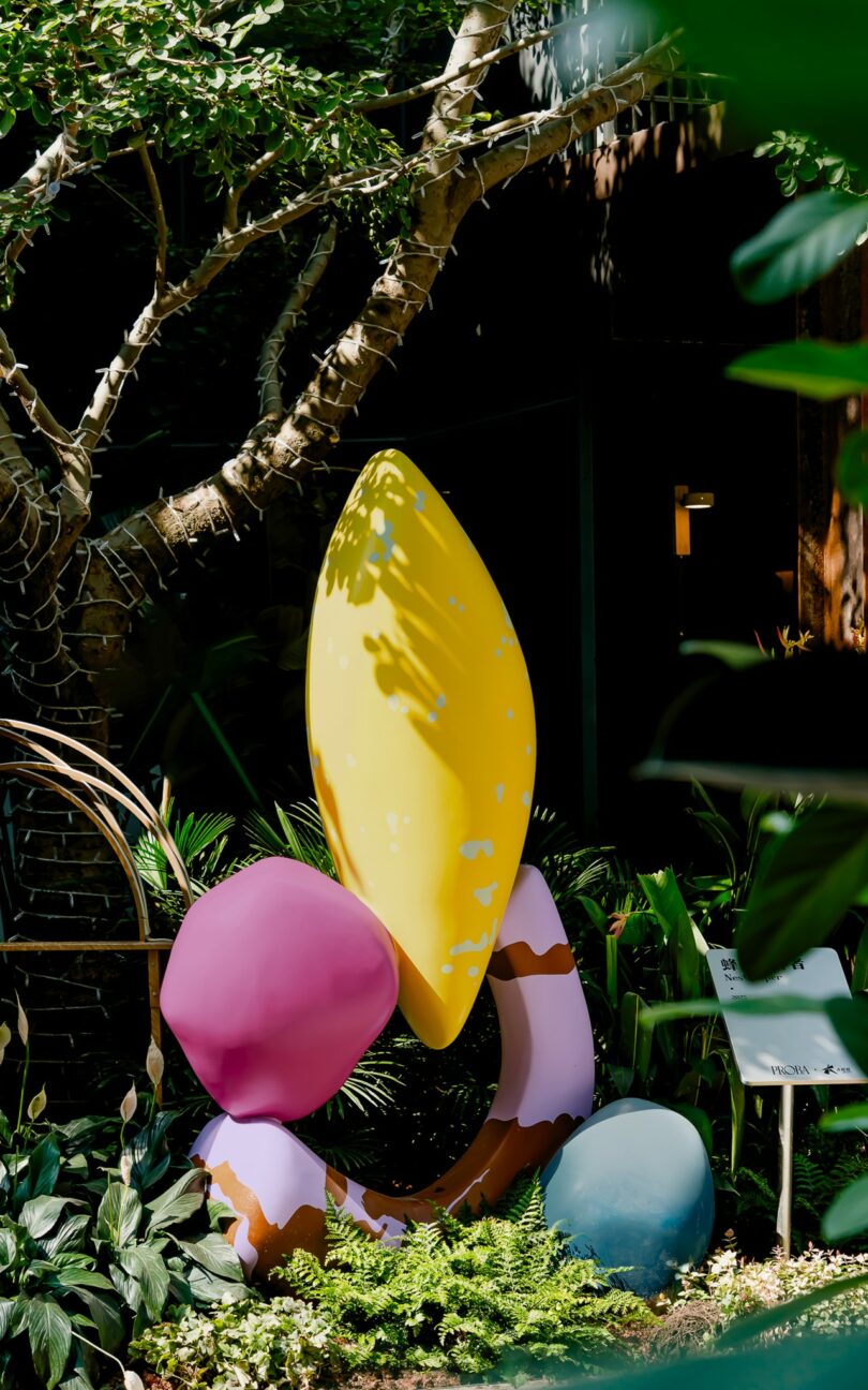 A colorful abstract sculpture by Studio Proba, with yellow, pink, brown, and blue shapes, stands among green plants and trees in an outdoor garden