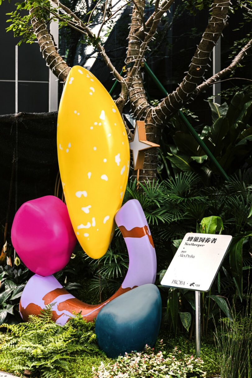 A large, colorful Studio Proba flower sculpture with pink petals and a green stem stands on a platform beside outdoor steps in an urban area with tall buildings and trees
