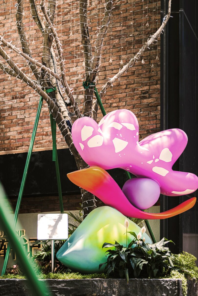 A colorful Studio Proba abstract sculpture with pink, purple, and green shapes sits in front of a leafless tree wrapped in string lights, set against a brick wall background