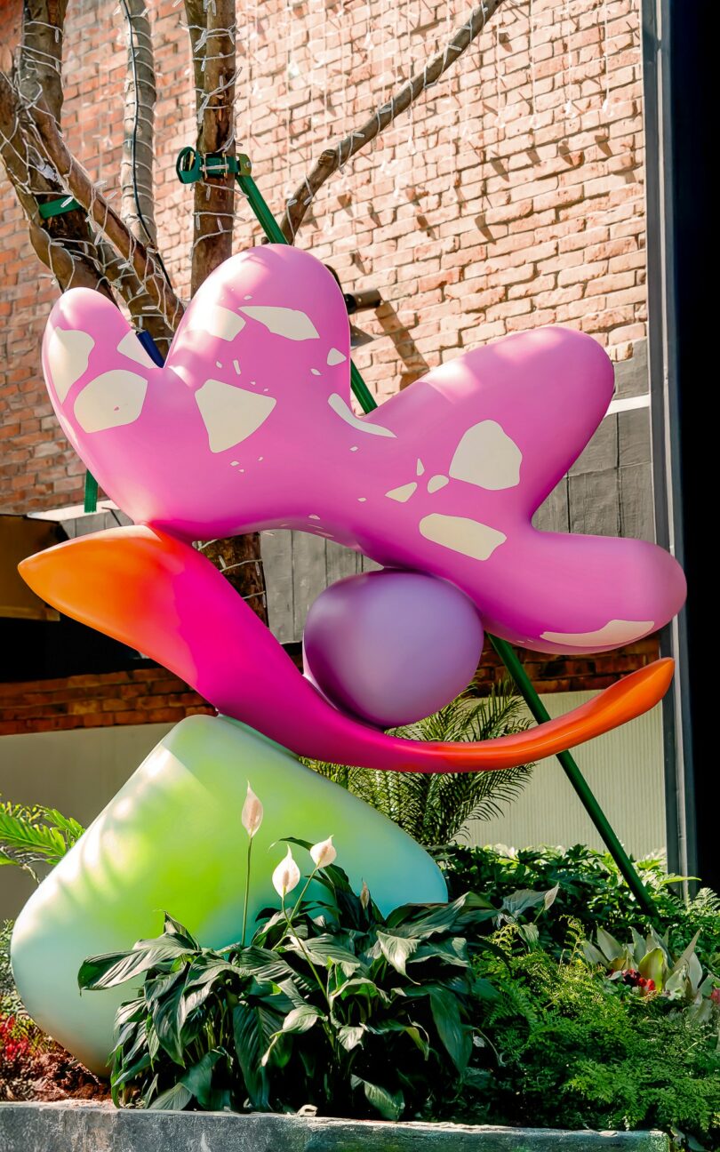 A colorful Studio Proba abstract sculpture with pink, purple, and green shapes sits in front of a leafless tree wrapped in string lights, set against a brick wall background