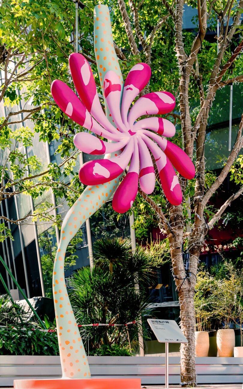 A large, colorful Studio Proba flower sculpture with pink petals and a green stem stands on a platform beside outdoor steps in an urban area with tall buildings and trees
