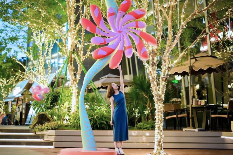 A woman in a blue dress stands outdoors, reaching up to touch a large, colorful, spiral-shaped Studio Proba sculpture surrounded by trees decorated with string lights