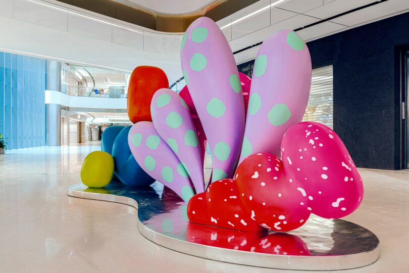 A colorful abstract sculpture with various organic shapes in pink, orange, yellow, purple, and green, displayed indoors on a shiny silver base