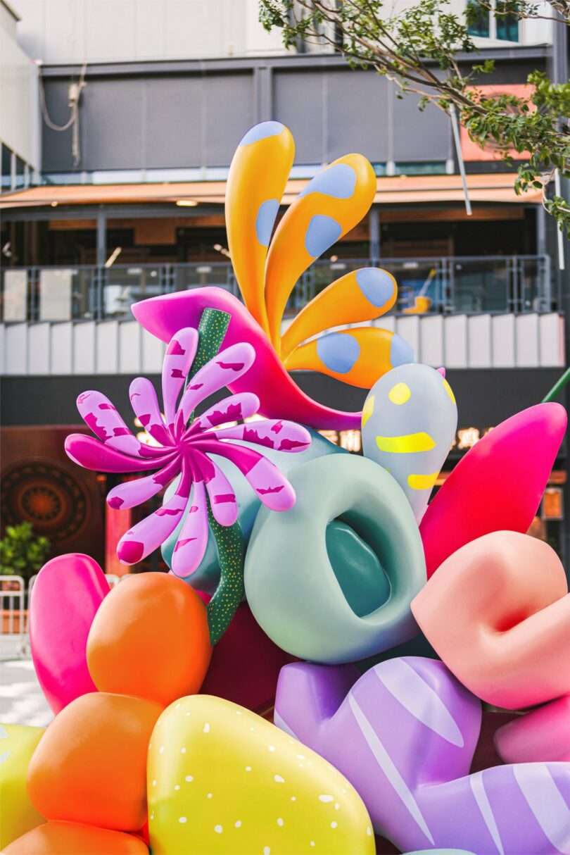 A colorful Studio Proba abstract sculpture with pink, purple, and green shapes sits in front of a leafless tree wrapped in string lights, set against a brick wall background