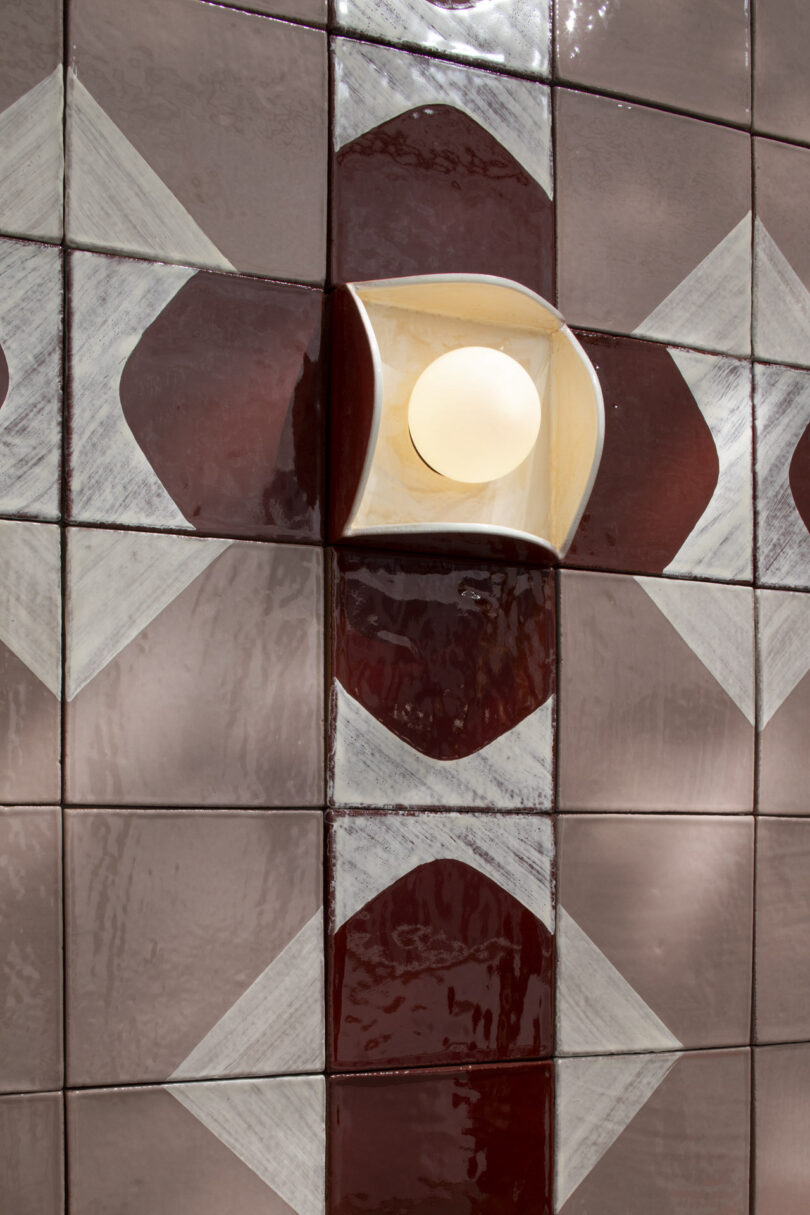 A round wall light fixture is mounted on a tiled wall with a geometric red and white diamond pattern