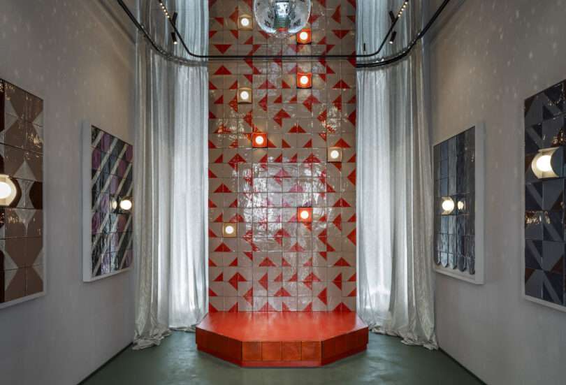 A geometric tiled wall with red and white triangles, red platform, mirrored disco ball above, and modern wall lights in a narrow room with gray curtains and framed artwork