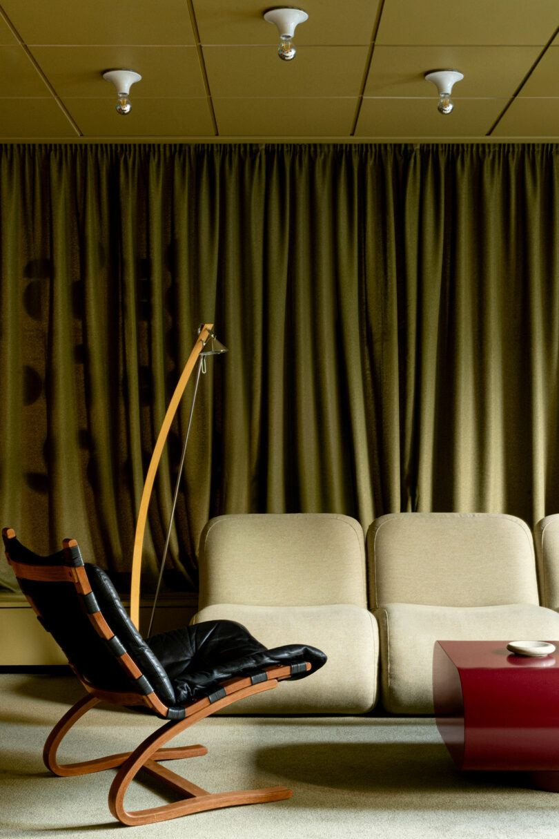 A modern living room with a beige sofa, a black leather lounge chair with wooden frame, a red coffee table, a floor lamp, and heavy green curtains.