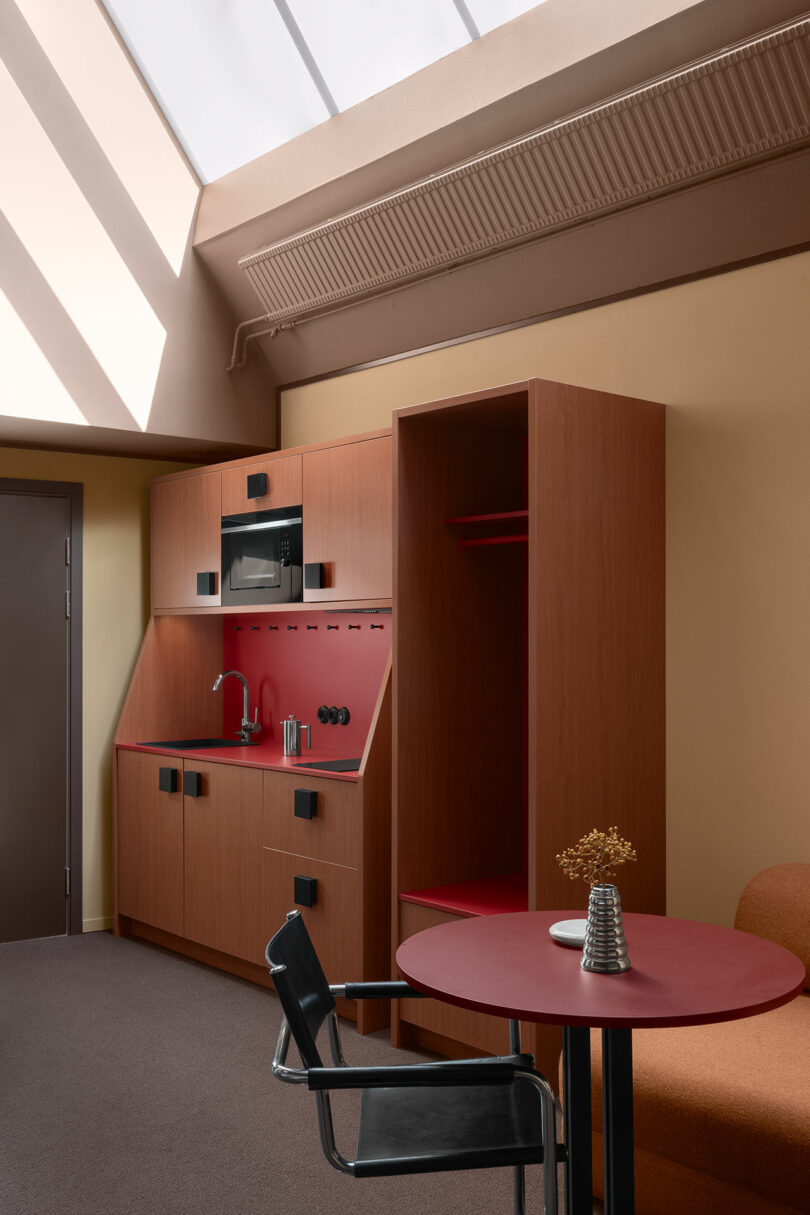A compact kitchenette with wooden cabinets, a sink, microwave, and open shelving next to a round table with a black chair and a small vase under a skylight.