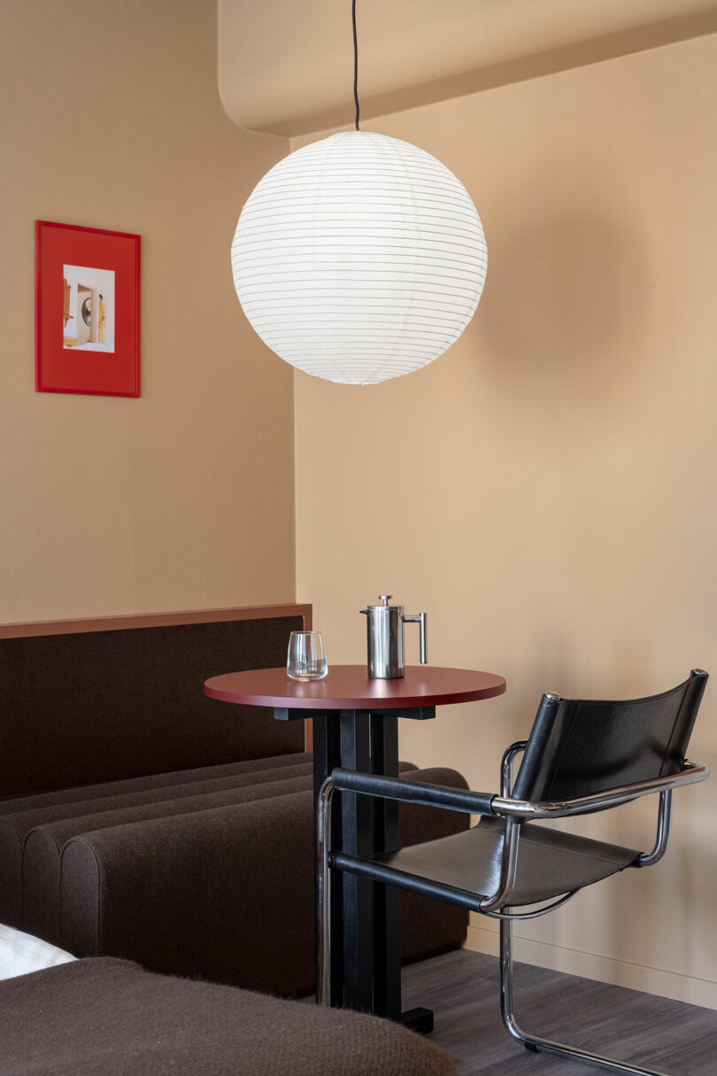 A small round table with a water glass, glass cup, and French press, next to a black chair under a hanging paper lantern in a modern, beige-walled room.