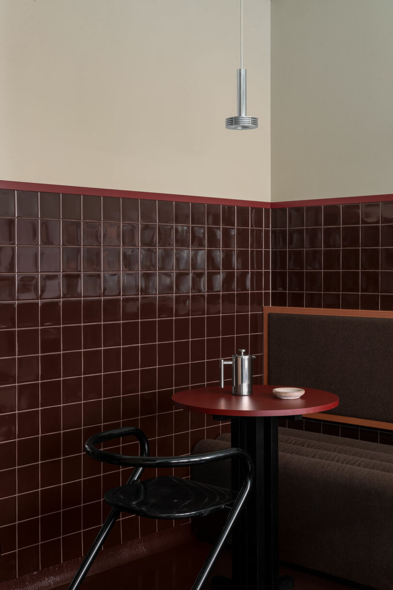 A cafe corner with dark brown tiled walls, a red table, a bench seat, a black high chair, a metallic coffee pot, a cup, and a pendant light overhead.