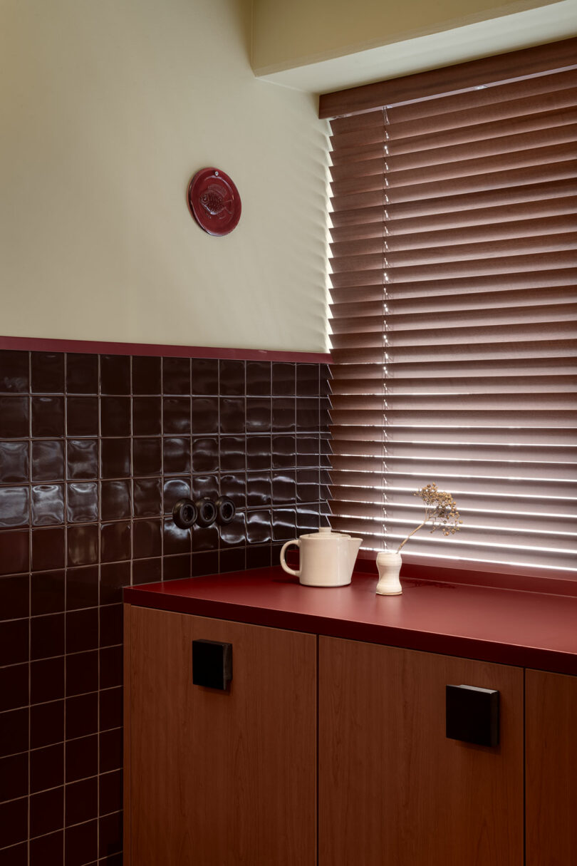 A kitchen corner with dark red tiled backsplash, red countertop, wooden cabinets, a white jug, a small vase with dried flowers, and closed brown blinds.