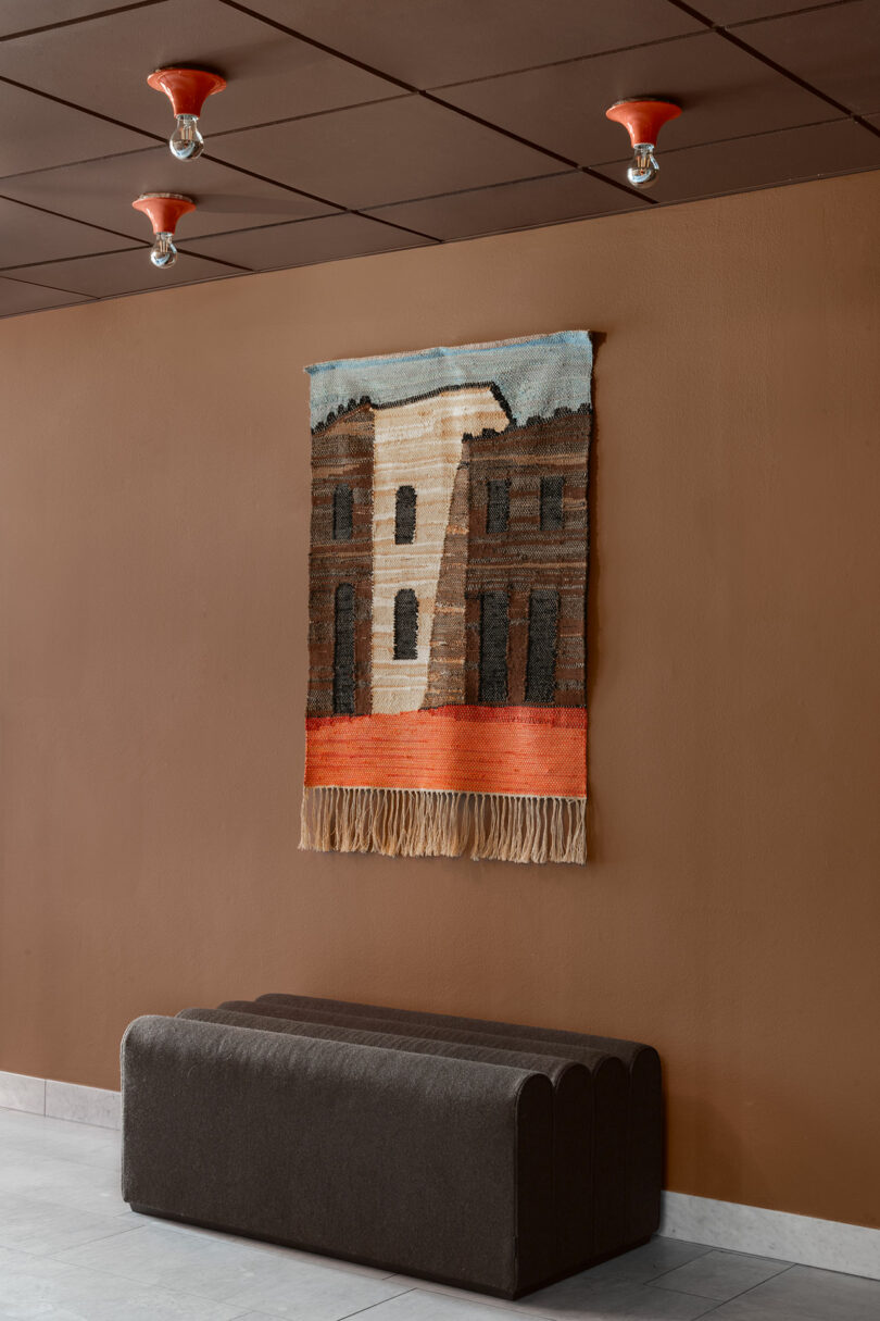 A woven wall tapestry depicting a building hangs on a brown wall above a dark upholstered bench; three red light fixtures are on the brown ceiling.