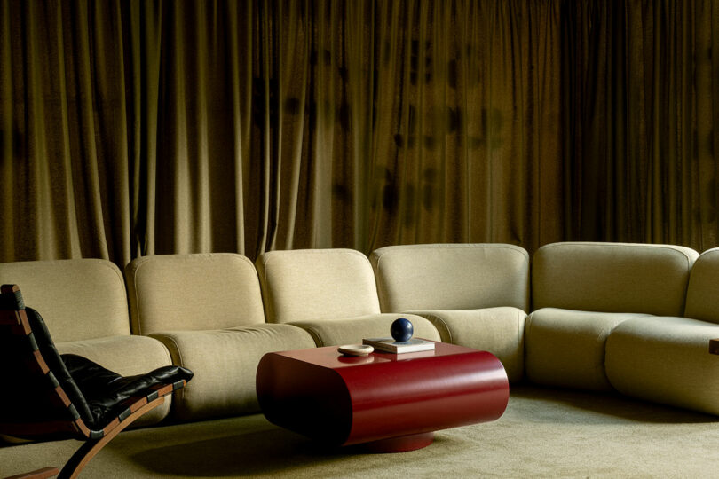 A modern living room with a beige curved sectional sofa, a black lounge chair, a red oval coffee table, and books, set against floor-to-ceiling brown curtains.
