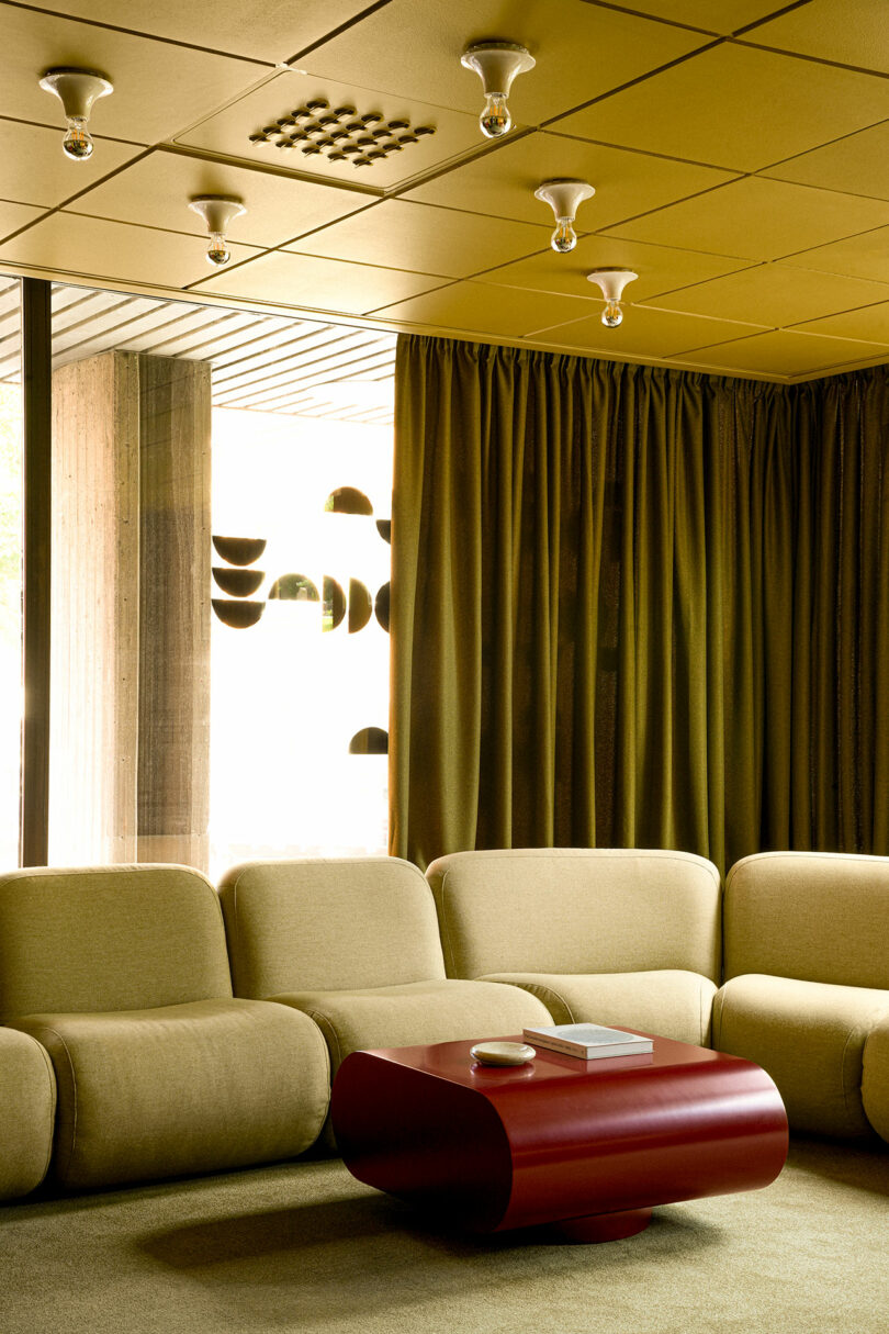 A modern living room with beige sectional sofa, red rectangular coffee table, olive green curtains, and exposed bulb ceiling lights.