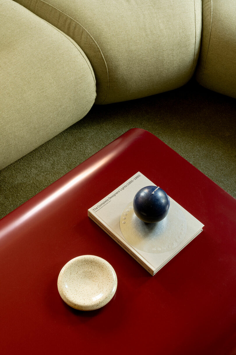 A red table with a beige bowl, a closed book, and a dark spherical object on top, in front of a light olive green sofa with curved cushions.