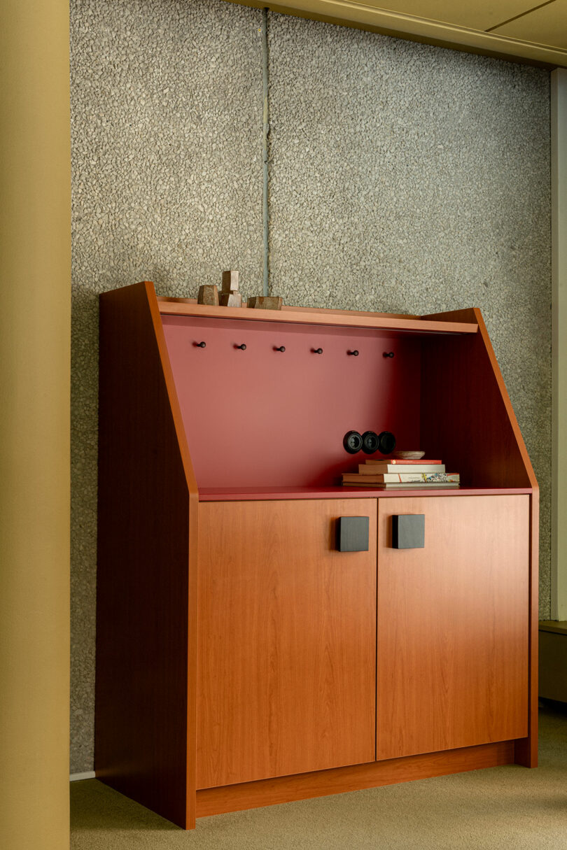 A wooden cabinet with a red back panel stands against a textured wall, holding books, small objects, and several electrical outlets on its shelf.